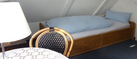 Una habitación pequeña con cama individual, techo inclinado, una silla de mimbre y una mesa con mantel de encaje.