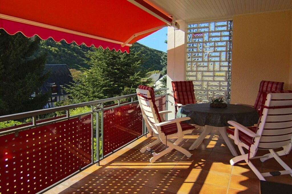 Un balcon doté d’un auvent rouge, de deux fauteuils à bascule blancs, d’une petite table ronde et offrant une vue sur la verdure et les bâtiments.