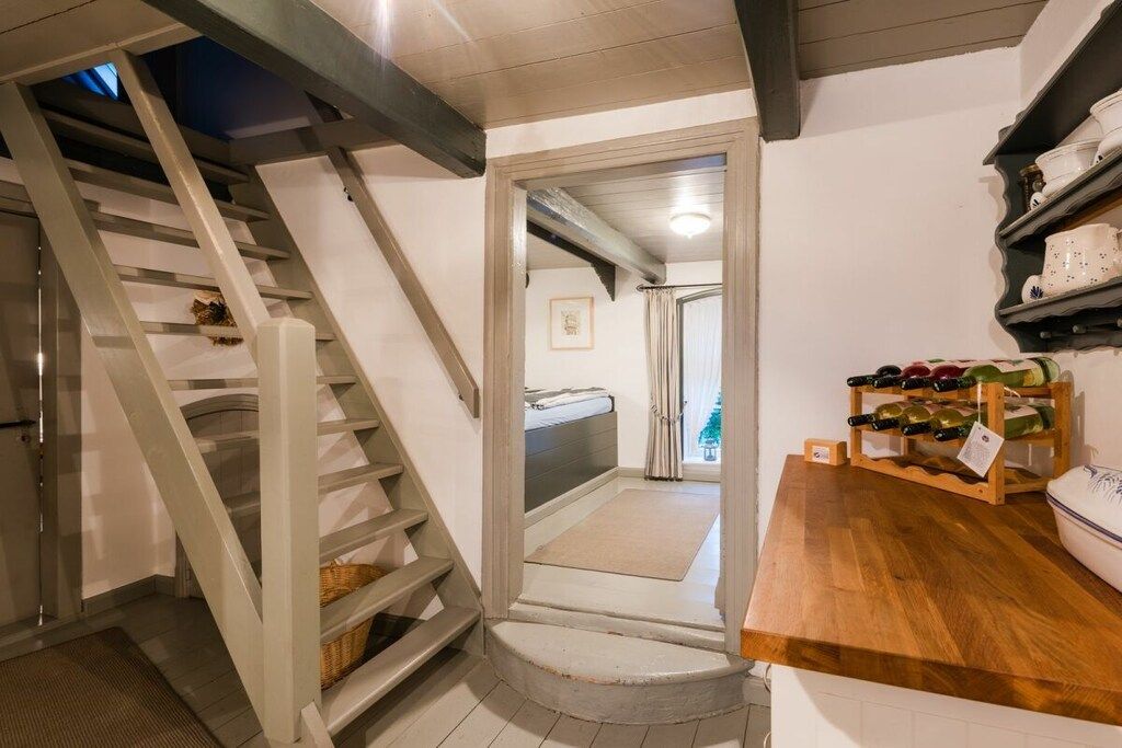Una escalera conduce a un dormitorio con cama y ventana. A la derecha, una estantería con botellas de vino.