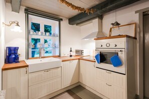 A kitchen with white cabinetry, a wooden countertop, a built-in oven, a microwave, and a window with decorative items. - Huus Campen - Vorderhuus (Campen)