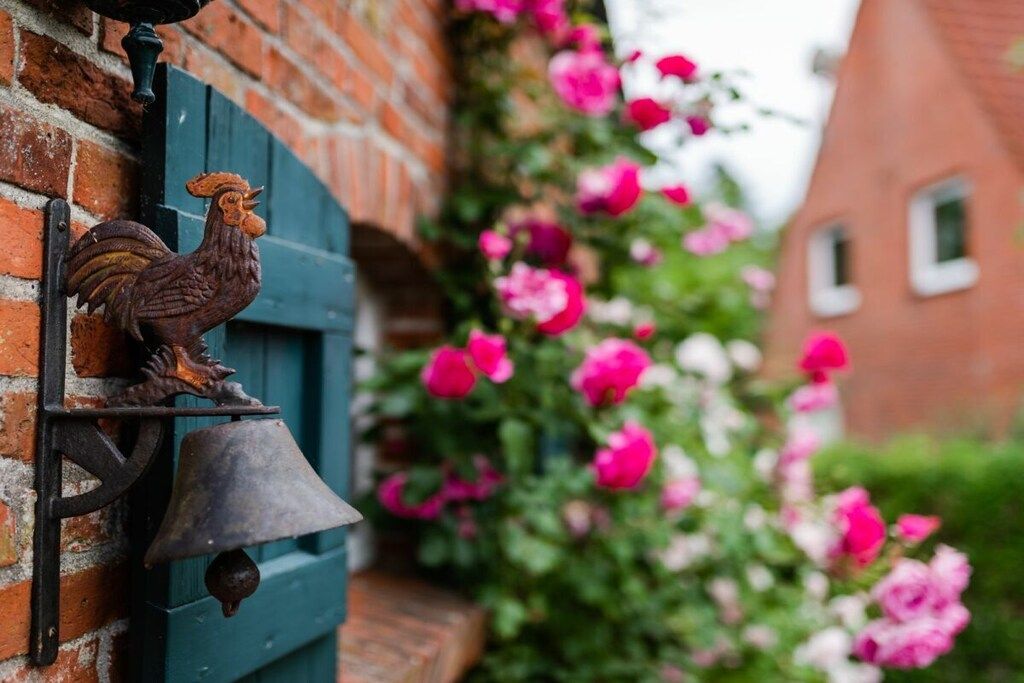 Ein Hahn als Wanddeko, eine Glocke und rosa Blumen in einem Garten.