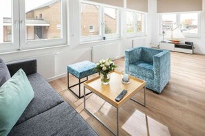 A modern living room with a sofa, a blue armchair, a wooden coffee table with a vase and remote control, and large windows with a view of buildings. - Holiday apartment in Juist with swimming pool (Juist)