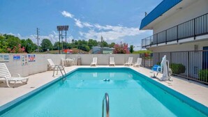 Una piscina rectangular con una escalera metálica en primer plano, rodeada de sillas de descanso blancas y un muro blanco con varios letreros.