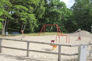 Ein Spielplatz mit Schaukeln und Rutsche, umgeben von Bäumen und einem Sandbereich.
