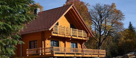 A wooden cabin with a balcony, situated on a rocky terrain with trees in the background.