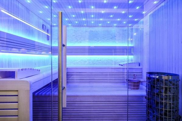Eine Sauna mit Glastür, Holzsitzen und einem Metallbehälter auf der rechten Seite.