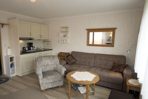 A compact living space with a kitchenette, a grey armchair, a brown sofa, and a small wooden table with a doily. - Apartment -Starfish- (Borkum)