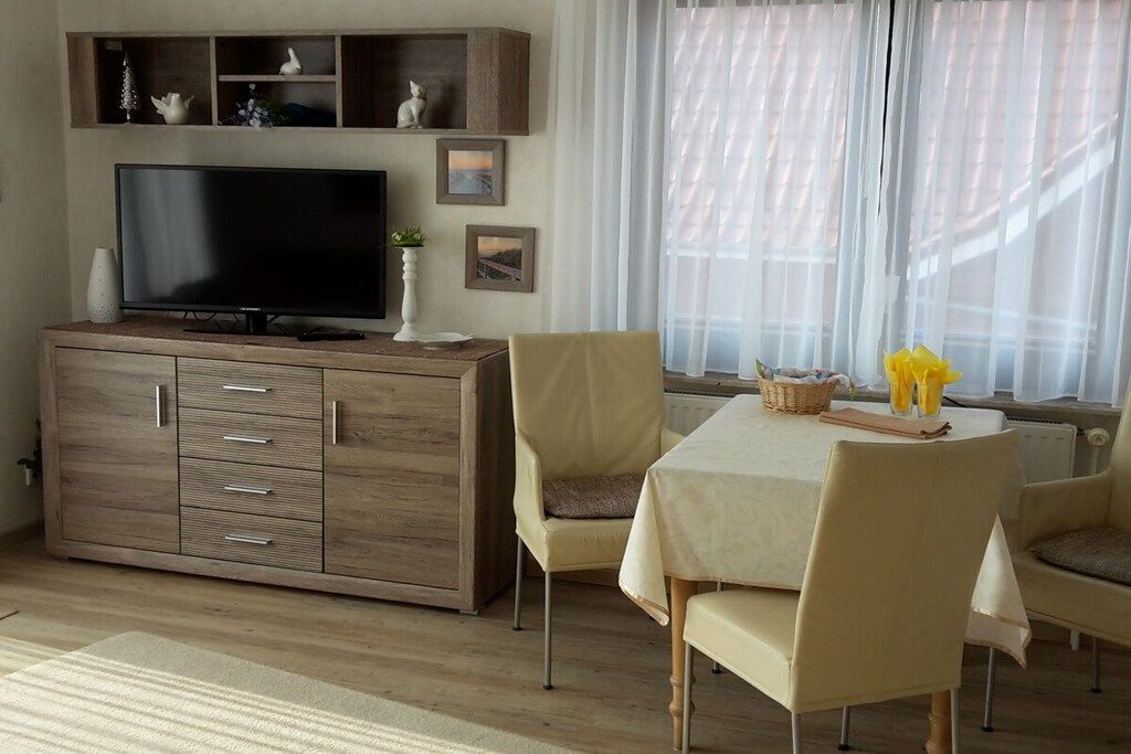 A living room with a flat-screen TV, wooden cabinet, beige chairs, a table with a white tablecloth, and a window with sheer curtains.