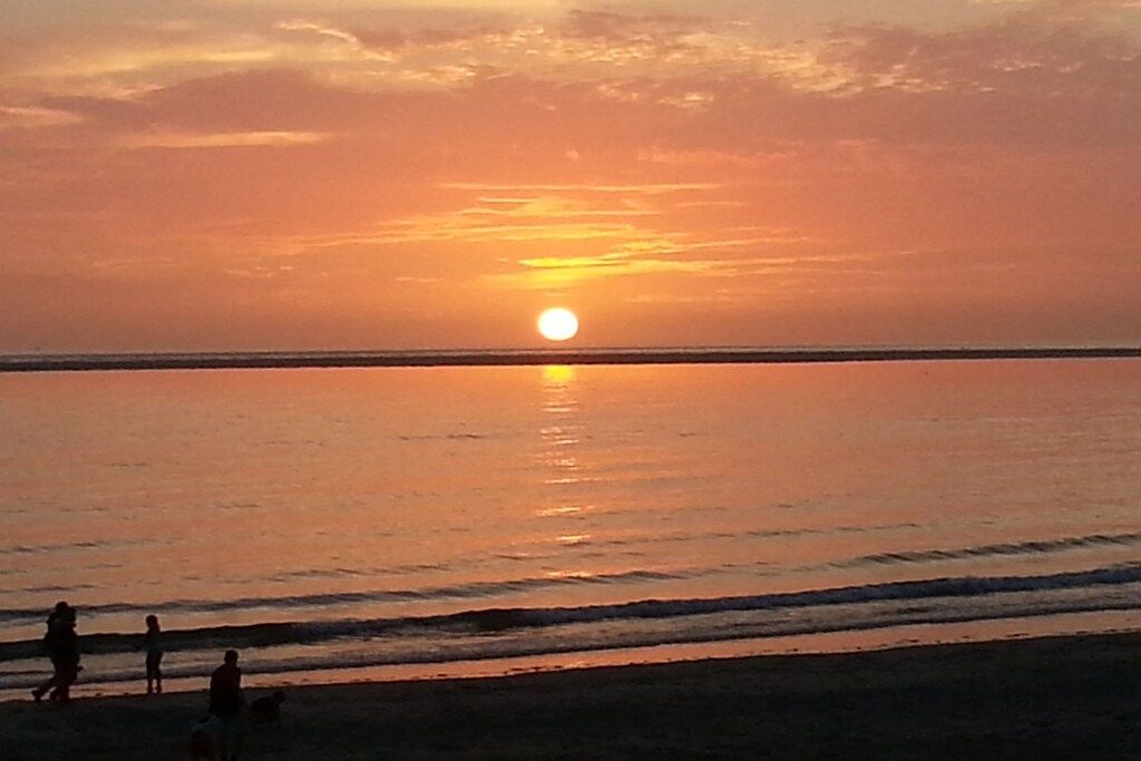 Pôr do sol sobre um mar calmo, com silhuetas de pessoas e um cão na praia.