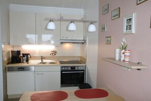 A compact kitchen with a dishwasher, oven, and a double sink. There are wall-mounted lights above the sink and a shelf with decorative items. - Residence Albatros 28 (Cuxhaven)