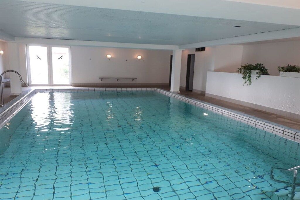 An indoor swimming pool with clear blue water, a tiled floor, and a bench along the wall.