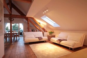 A loft-style room with a sloped ceiling, wooden beams, a skylight, a white sofa, a dining table, and a rug. - Castle view at Sonnenbrink (Wernigerode)