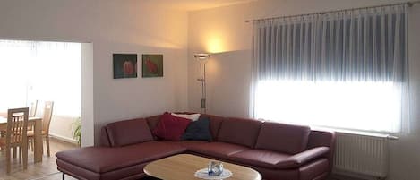 Una sala de estar con un sofá modular, una mesa de centro, un comedor con sillas y un ventanal con cortinas.
