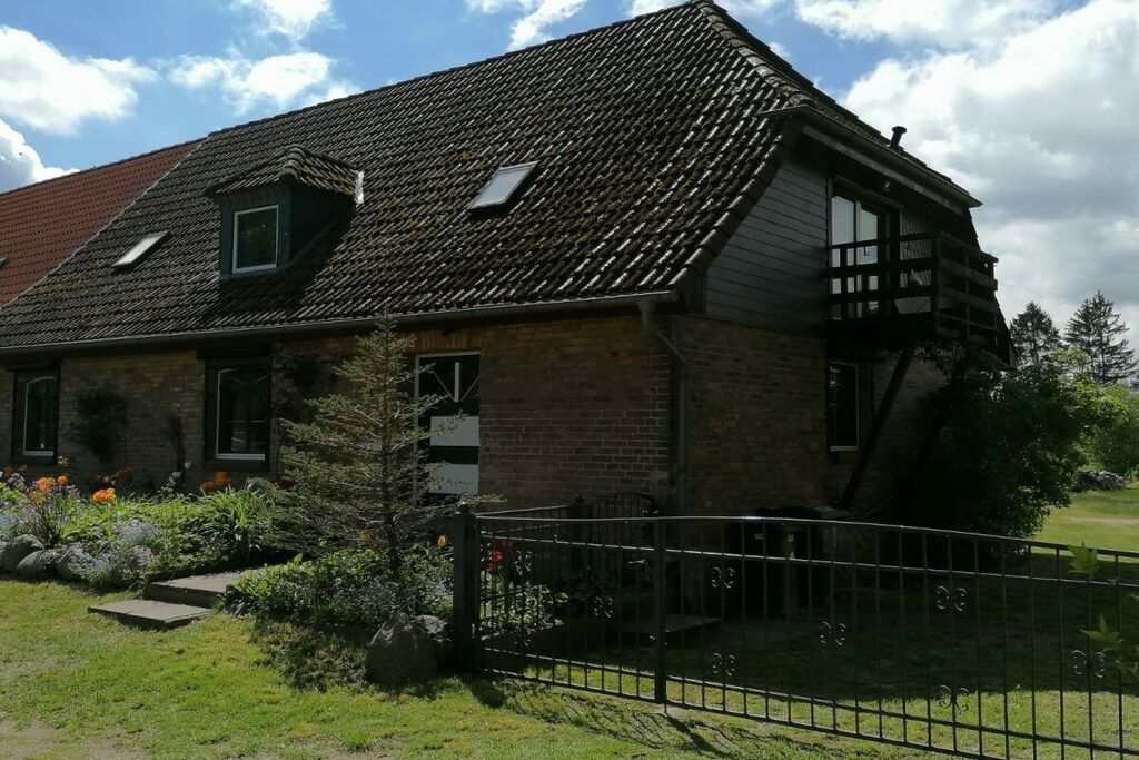 Une maison à deux étages avec un toit en pente, un petit garage attenant et une cour clôturée où poussent différentes espèces de plantes.