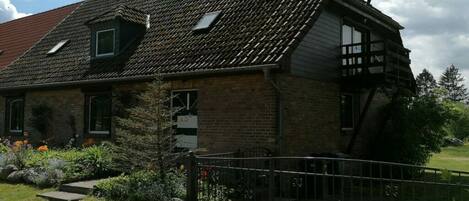 Une maison à deux étages avec un toit en pente, un petit garage attenant et une cour clôturée où poussent différentes espèces de plantes.