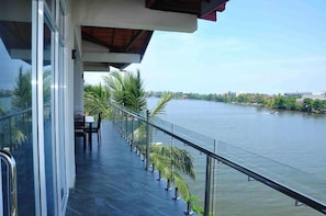 A balcony with a glass railing overlooking a calm body of water, palm trees, and distant buildings.