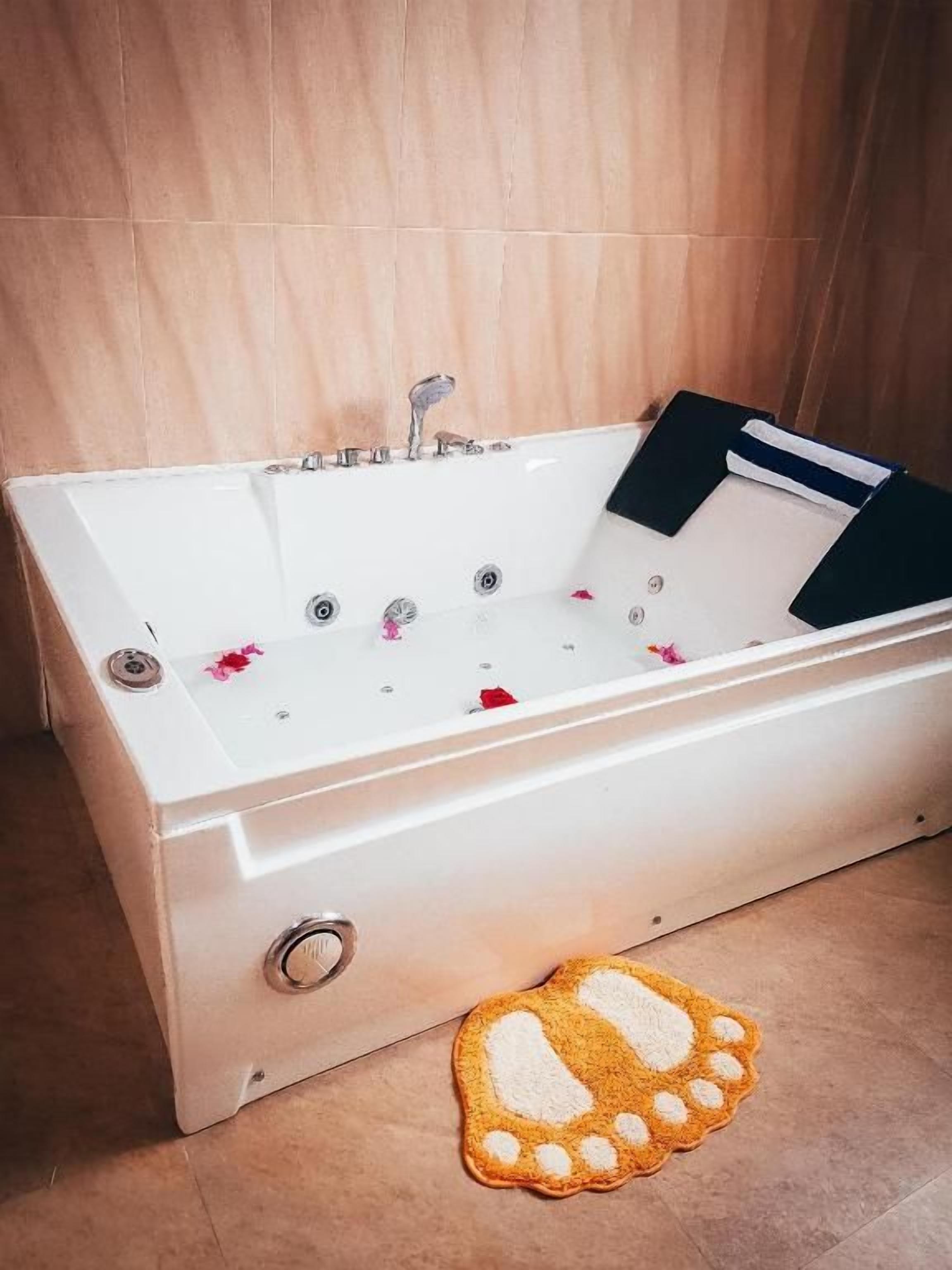 A white bathtub with multiple faucets, a black towel, and a pair of slippers on the floor.