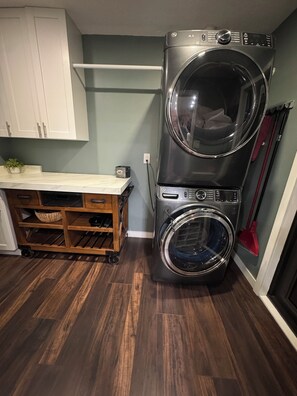 A compact laundry room with a stacked washer and dryer, wooden storage unit, and a red broom. - Spacious 4-bedroom house in peaceful Delray Beach minutes from Atlantic Ave! (Delray Beach)