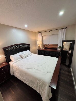 A neatly made bed with white linens, a wooden headboard, a bedside table with a lamp, and a dresser with a mirror. - Spacious 4-bedroom house in peaceful Delray Beach minutes from Atlantic Ave! (Delray Beach)