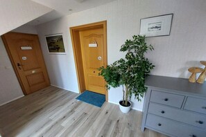 Een gang met twee houten deuren, een bloempot, een lijst met een foto aan de muur en een grijze dressoir.