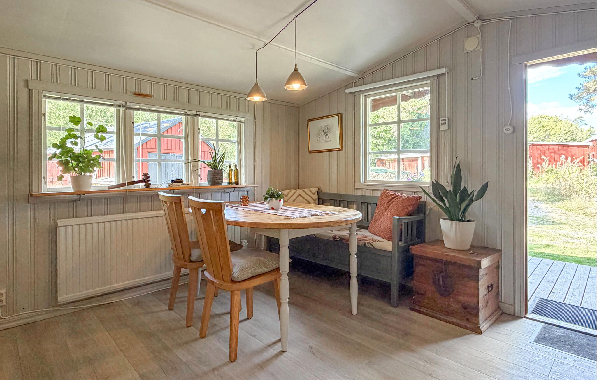 A cozy kitchen with wooden chairs, a round table, and a bench. There are windows with plants and a view of the outside.
