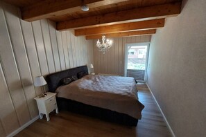 A bedroom with a wooden ceiling, a bed with a dark headboard, a bedside table with a lamp, and a chandelier. - Holiday apartment with 1 bedroom (Butjadingen)