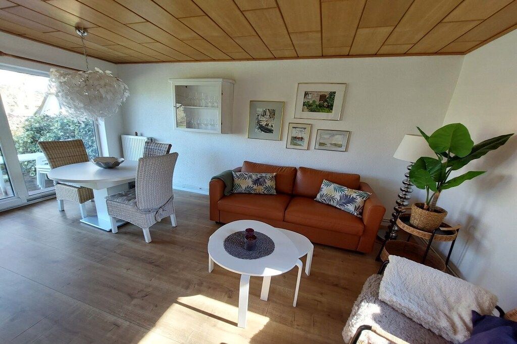 Un salón con techo de madera, una mesa redonda de comedor con sillas, un sofá con cojines, una mesita blanca con un objeto decorativo y una planta en maceta.