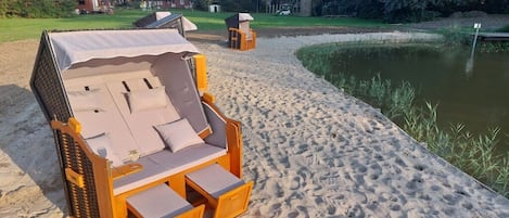 A beachside wooden lounge chair with a canopy, positioned on a sandy area adjacent to a pond.