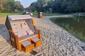 En solstol i trä med ett parasoll, placerad på en sandstrand intill en damm.