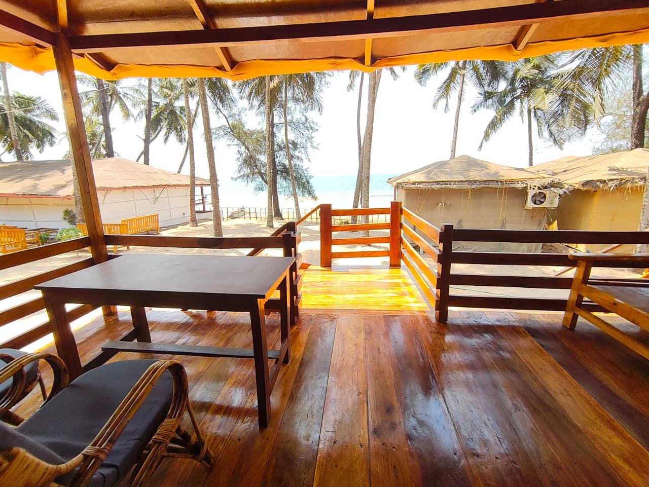 An outdoor wooden deck with tables and chairs, overlooking a beach with palm trees.
