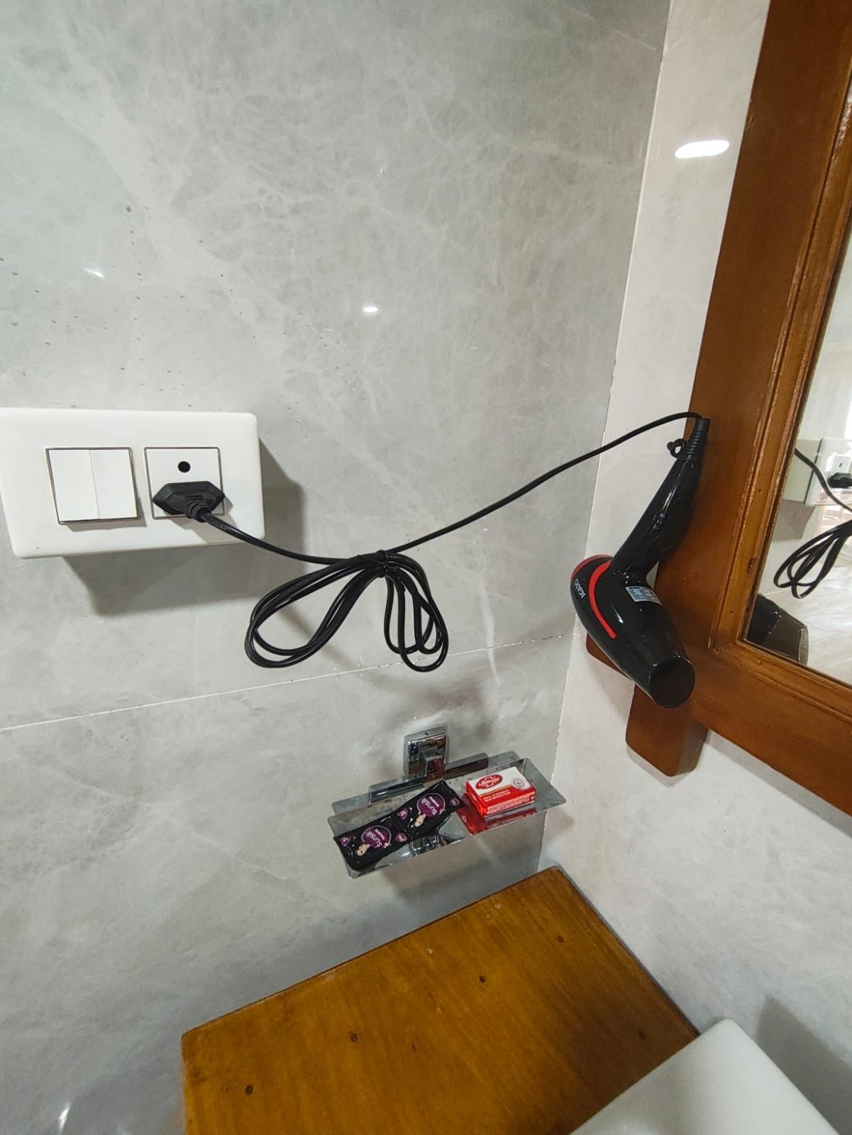 Un baño con pared de azulejos blancos, un enchufe, un secador de pelo y un pequeño estante con artículos de aseo.