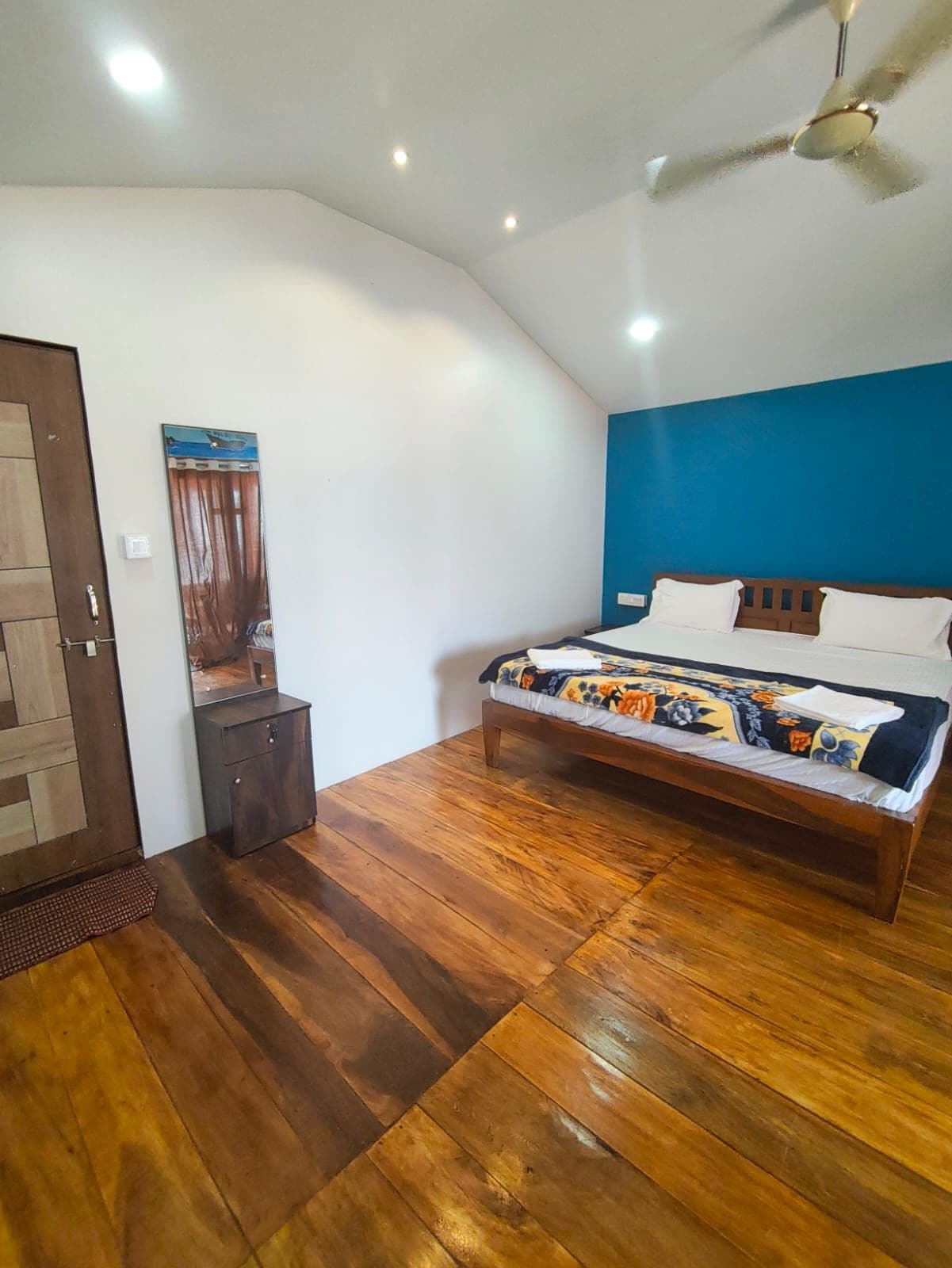 A bedroom with a wooden floor, a bed with a floral bedspread, a ceiling fan, and a door with a wooden panel design.