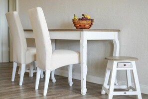 Ein Essbereich mit einem weißen Tisch und Stühlen, einem kleinen Hocker und einer Obstschale auf dem Tisch.