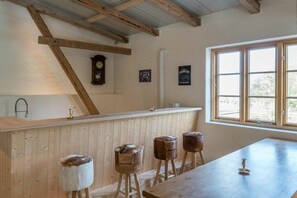 A rustic interior with a wooden bar, three stools with fur seat cushions, a wooden ceiling with exposed beams, a window with a view, and a wall-mounted clock. - Kohstall in the Haubarg Ossenhoff (Tating)
