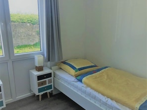 A single bed with a yellow blanket and pillows, a bedside table with a lamp, and a window with curtains. - House No. 20, Fischerhus (Wangerland)
