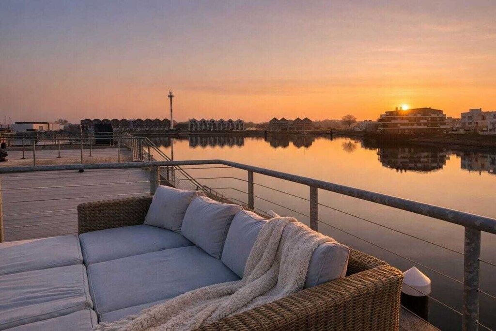 A waterfront balcony with a wicker sofa and a view of the sunset over a calm body of water.
