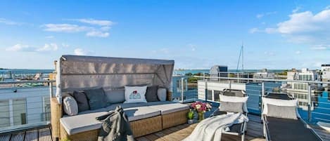 A sunlit deck with a sofa, cushions, a small table with drinks, and a view of the sea and buildings.