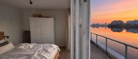 A bedroom with a bed, a wardrobe, and a view of a calm body of water during sunset.