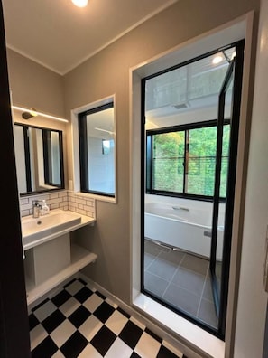 Un baño con piso a cuadros blanco y negro, un lavamanos blanco y un amplio ventanal con vista a la vegetación.