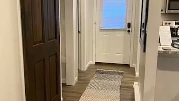 Un pasillo con una puerta de madera oscura, una puerta blanca y una zona de cocina con microondas.
