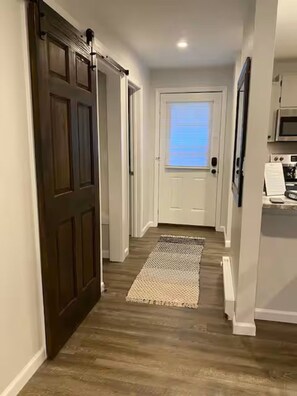Un pasillo con una puerta de madera oscura, una puerta blanca y una zona de cocina con microondas.