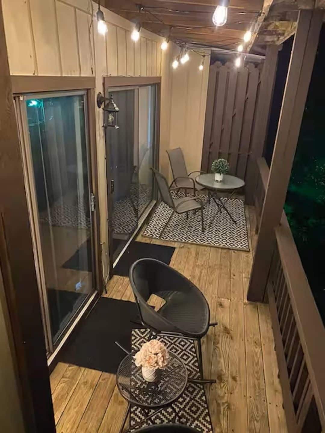 Un espacio exterior cubierto con terraza de madera, puerta de vidrio que da a una habitación, una pequeña mesa redonda con sillas y una planta en maceta.