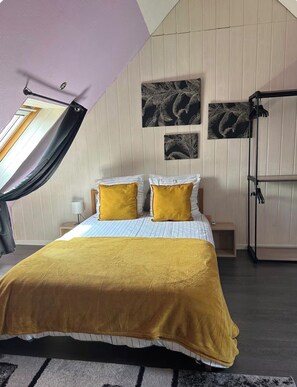 Ein Schlafzimmer mit schrägem Dach, einem Bett mit einer senffarbenen Decke, zwei Kissen, einem Nachttisch, einer Lampe und Wandkunst.
