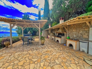An outdoor patio with stone flooring, a covered seating area, and a stone wall with a built-in grill. - Villa Artemis, Tolo, Nafplio (Tolo)