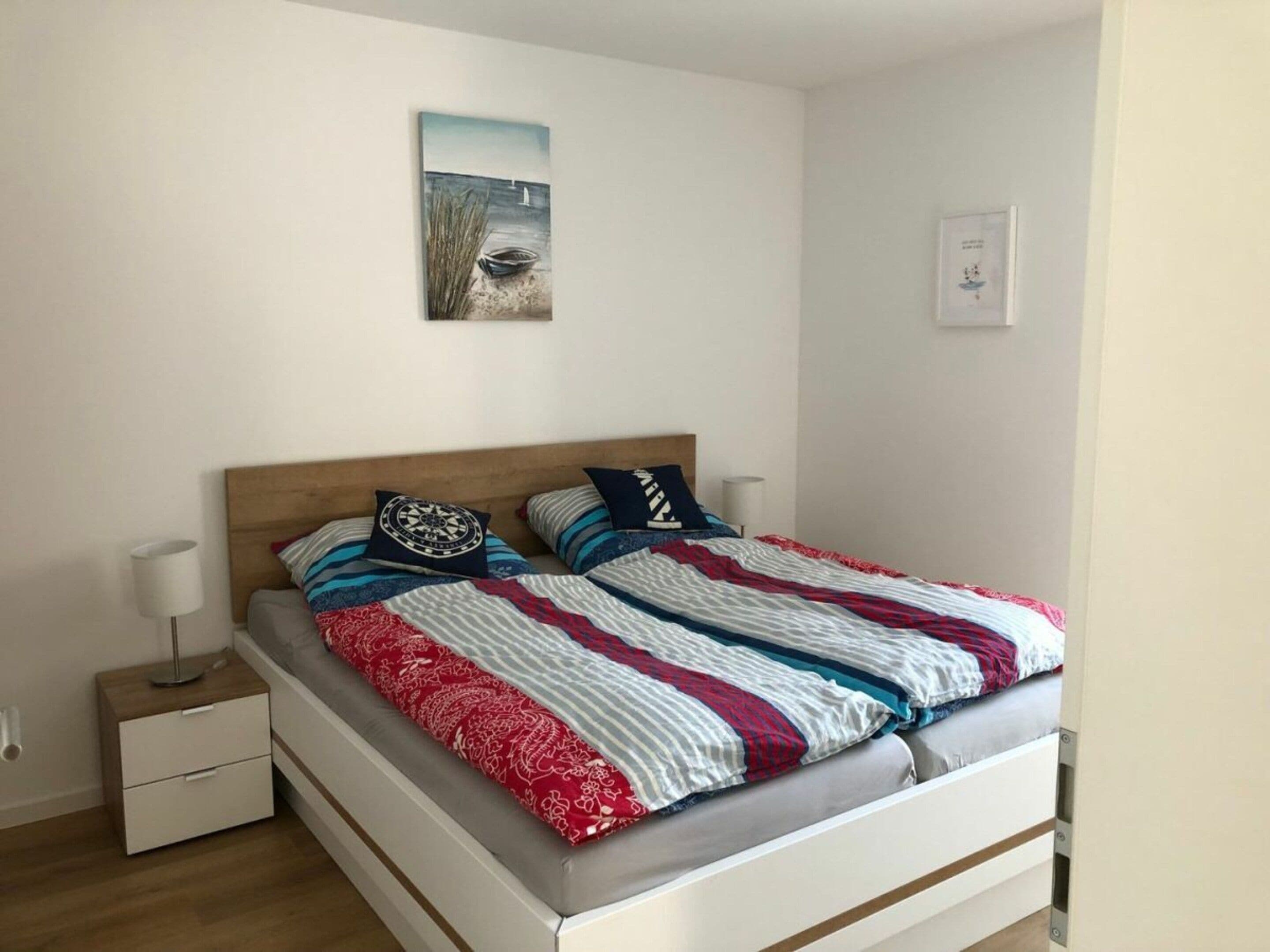 Ein ordentlich bezogenes Bett mit einem gestreiften Überwurf und Kissen, ein Nachttisch mit einer Lampe und ein Wandbild.