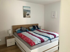 A neatly made bed with a striped blanket and pillows, a bedside table with a lamp, and a wall-mounted picture. - 50.1.n North Bunk (Wangerland)