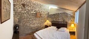 A room with a stone wall, a bed with white linens, two bedside lamps, and a fan. - MARTINE'S HOUSE (La couarde sur mer)