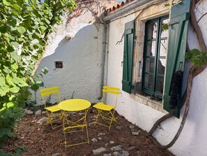 A small outdoor seating area with a table and two chairs, a window with green shutters, and a white wall. - MARTINE'S HOUSE (La couarde sur mer)