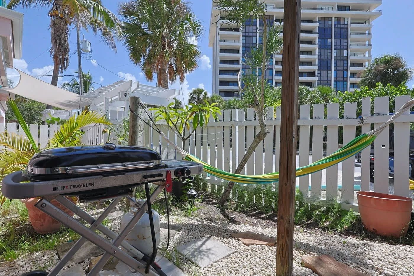 A portable grill with a metal frame and wheels, positioned in a garden area with a white picket fence, palm trees, and a building in the background.