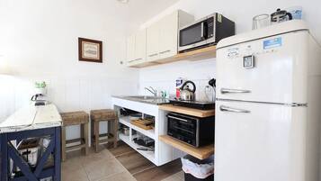 A compact kitchen with a microwave, oven, and refrigerator. There are two stools and a small table with a kettle.